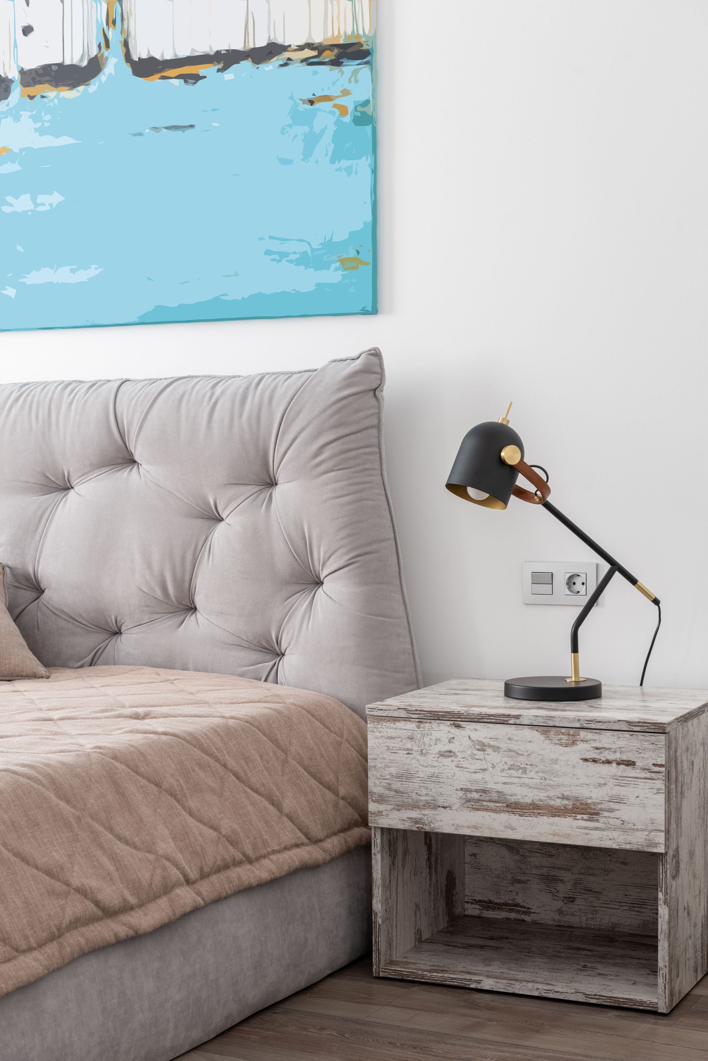 Interior of modern light hotel room with bed with blanket near table with lamp and painting on wall