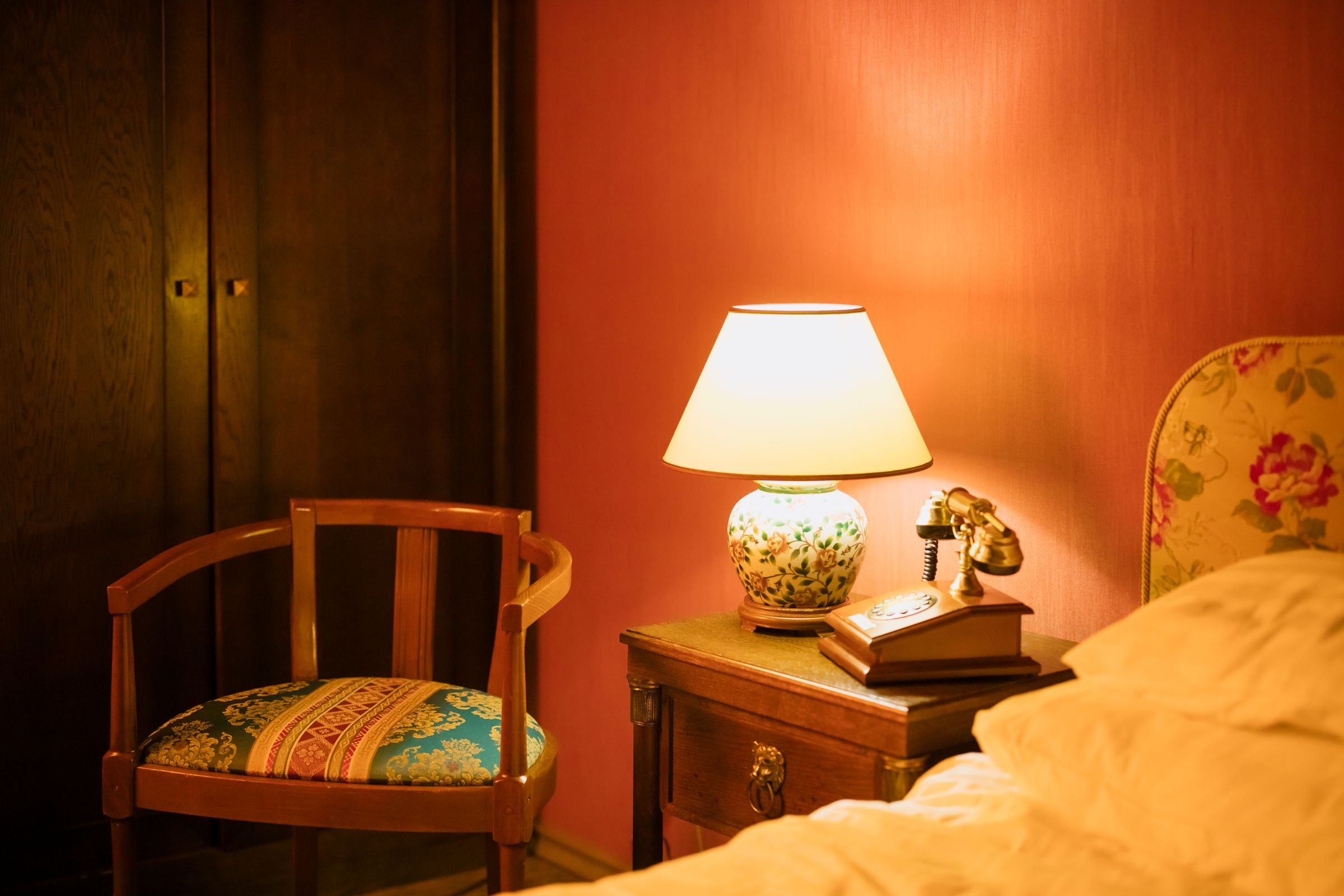 Warm vintage bedroom interior featuring a lamp, chair, and telephone by the bedside.