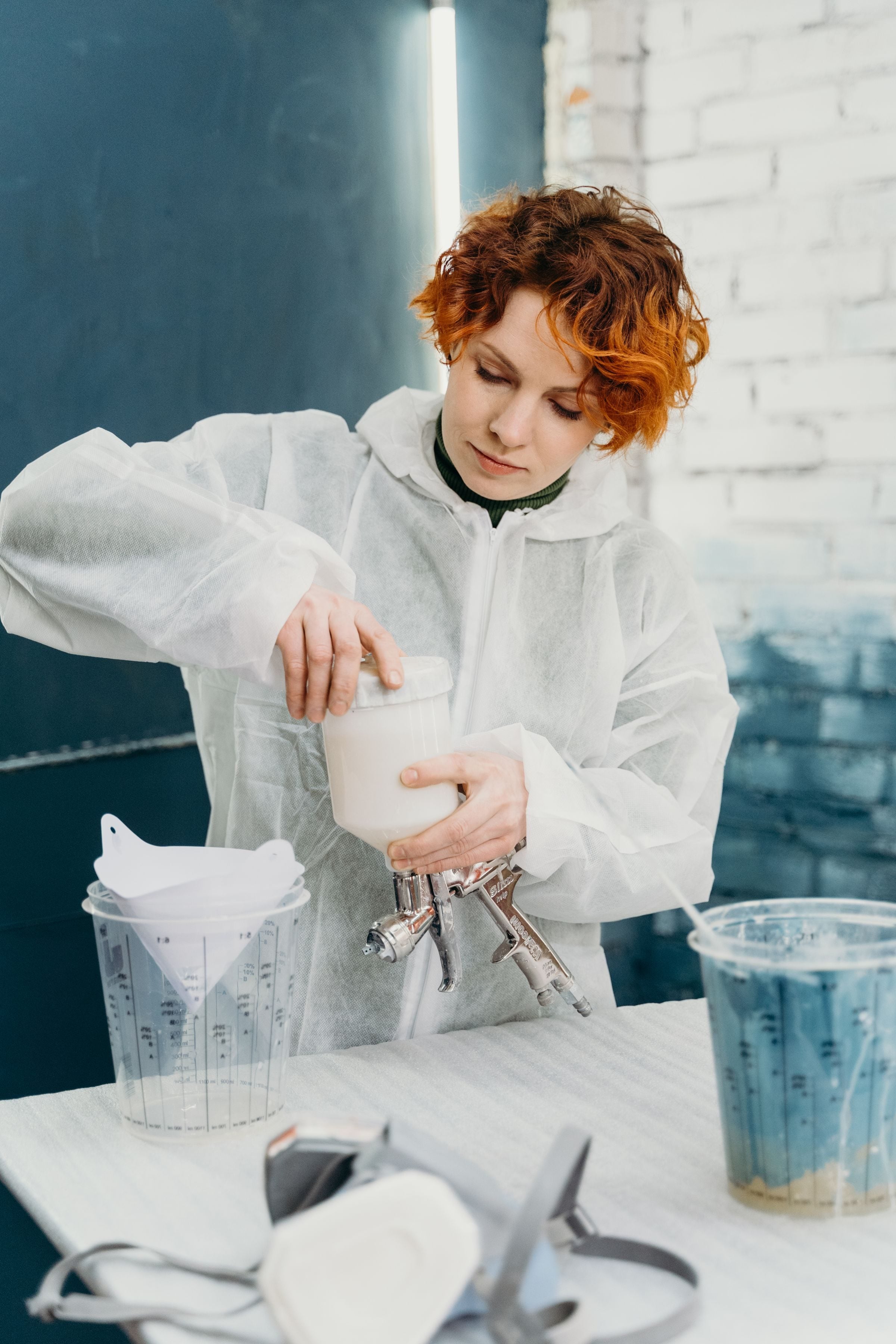 Redheaded woman in protective suit preparing spray paint in workshop.