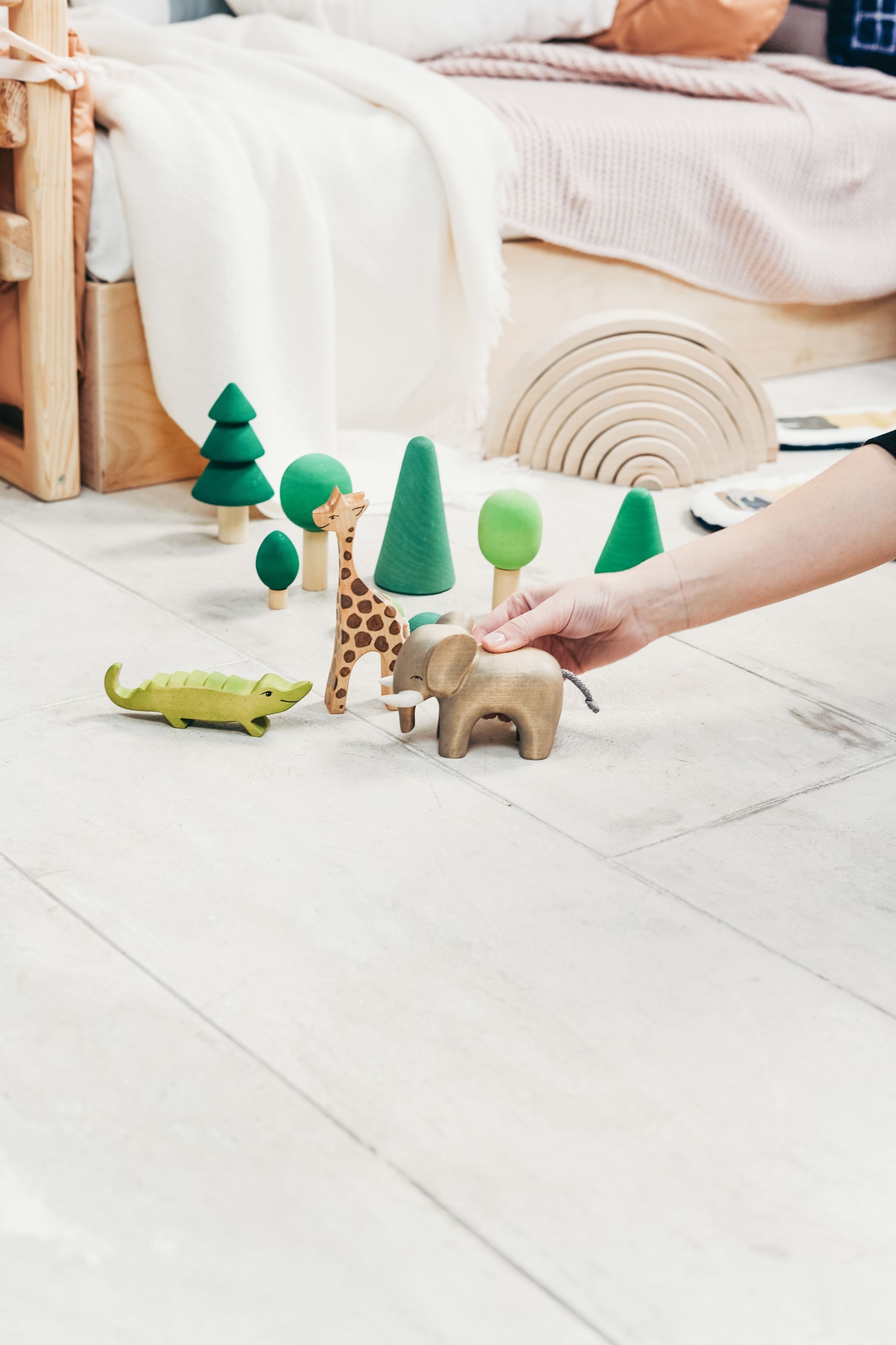 A playful indoor scene featuring a child's hand engaging with wooden toy animals and trees.