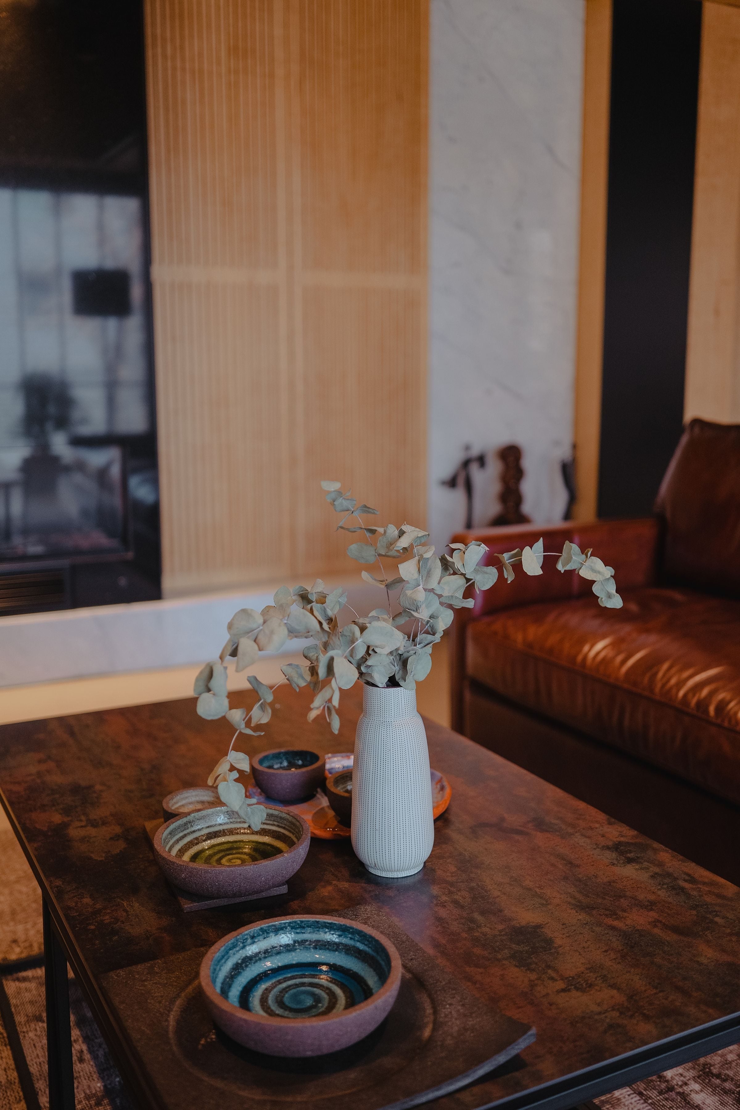 A modern living room with a designer coffee table, decorative plates, and a vase with eucalyptus, featuring warm, natural tones.